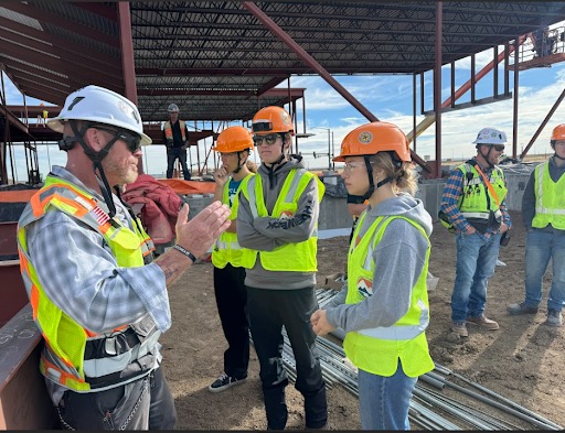 A construction worker in a hard hat and safety vest speaks to a small group of people, including two young people, on a construction site with exposed steel framing in the background.