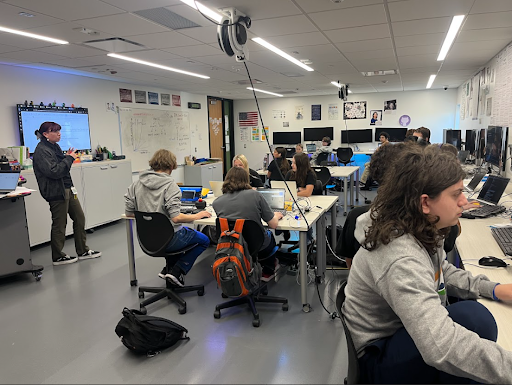 A wide shot of a brightly lit computer science classroom with a teacher standing near a large screen and students working on laptops at tables and at rows of desktop computers.