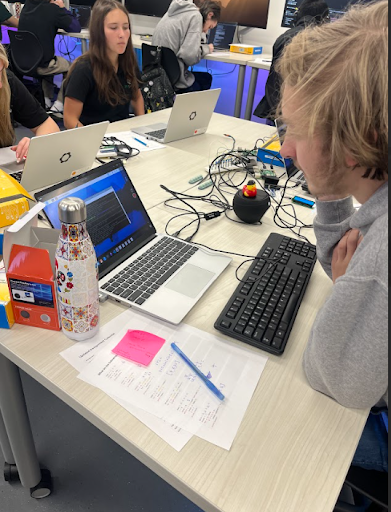 A student leans over a table with a laptop displaying code, a keyboard, a water bottle, and papers, surrounded by wires and other students in a computer lab.