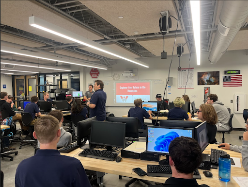 A wide shot of a computer lab where two presenters are talking to a large group of students seated at computers, with a presentation slide visible on the back wall.
