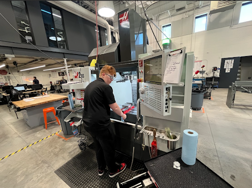 A young man in a black shirt and safety glasses operates a large CNC milling machine in a workshop setting.