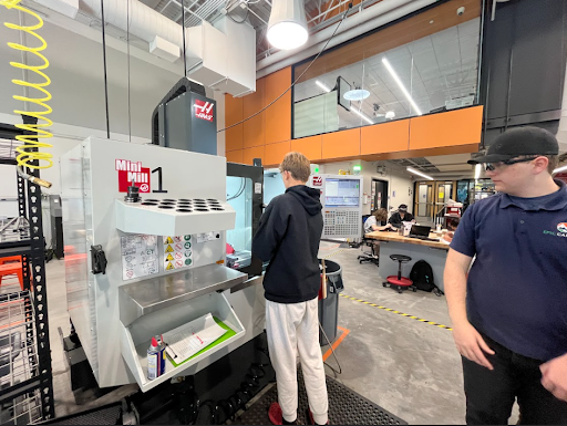 Two young men stand near a large HAAS Mini Mill CNC machine in a brightly lit industrial classroom or workshop.