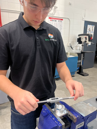 A close-up of a student wearing safety glasses and an EPIC CAMPUS shirt, using a tap handle to thread a metal piece held in a vise.