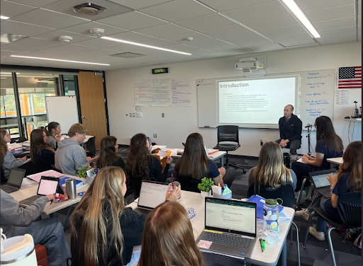 A presenter sits in front of a white screen displaying the word "Introduction" while speaking to a diverse class of high school students seated at desks with laptops in a bright, modern classroom.