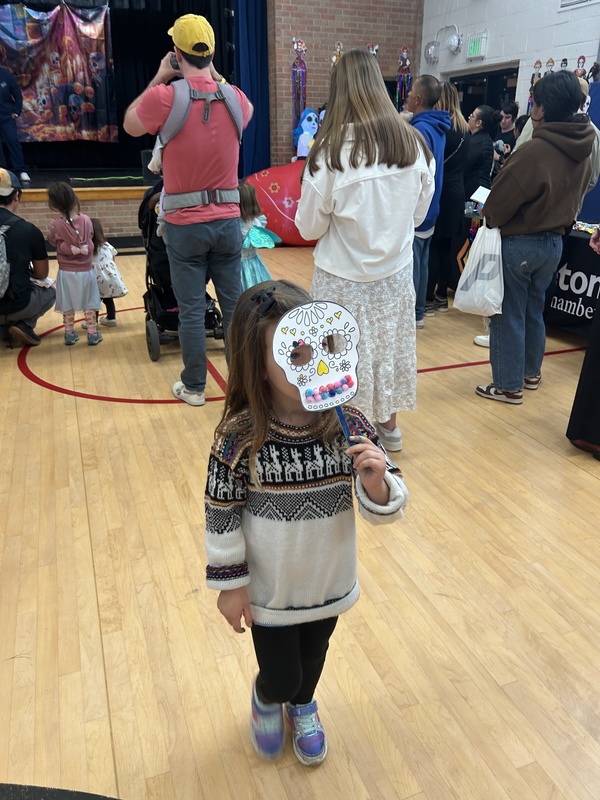 A young girl in a sweater holds a decorated paper sugar skull mask at a Día de Muertos event.