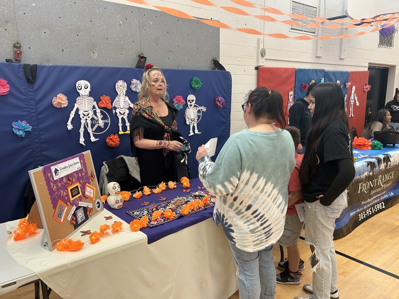 An adult with face paint stands behind a vendor table decorated with paper skeletons and marigolds, talking to two females.