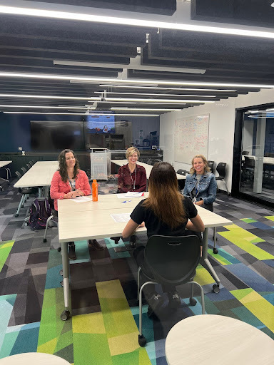Four people sitting at a table in a modern office space, three facing the camera and one with their back to it, appearing to be conducting an interview or meeting.