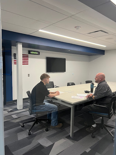 Two men sitting across from each other at a large conference table in a room with a TV screen and an American flag on the wall. They appear to be in a meeting or interview.