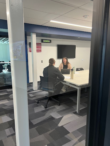 A woman and a person in black sitting across a table from each other in a conference room, visible through glass. The woman is using a laptop.