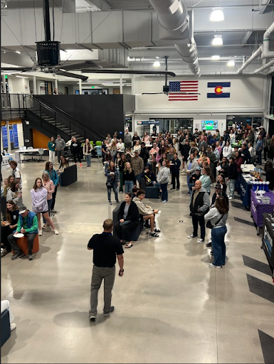 A large crowd of students and people attending an event in a spacious, two-story indoor area with a speaker addressing them in the foreground. US and Colorado flags hang overhead.