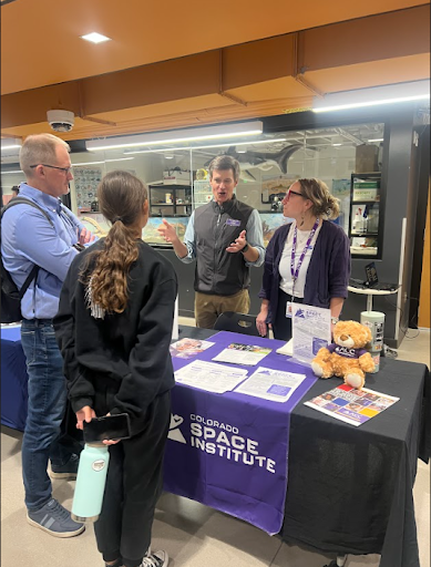 Four people gathered around a table with a Colorado Space Institute banner. A man is gesturing while speaking to a student and another adult.