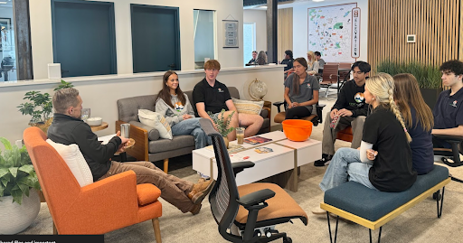A group of students and an adult sitting in a modern, casual office space, engaging in a relaxed discussion or mentorship session.