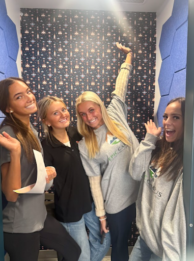 Four young women smiling and posing playfully inside a small booth with space-themed wallpaper and blue acoustic panels.