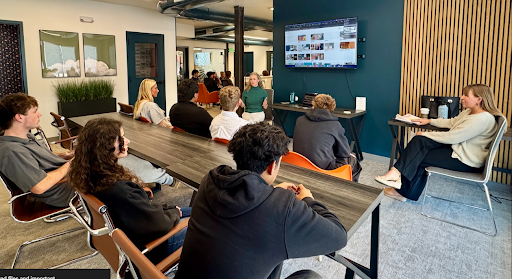 A group of students seated around a long table listening to a presentation given by two women in a modern office or classroom.