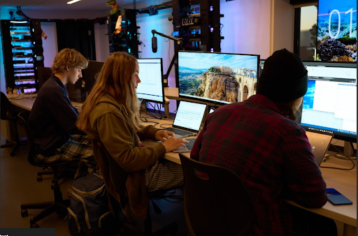 Three students sitting at computers in a dimly lit, high-tech room, working on laptops and desktop monitors, possibly during a CyberPatriot competition.