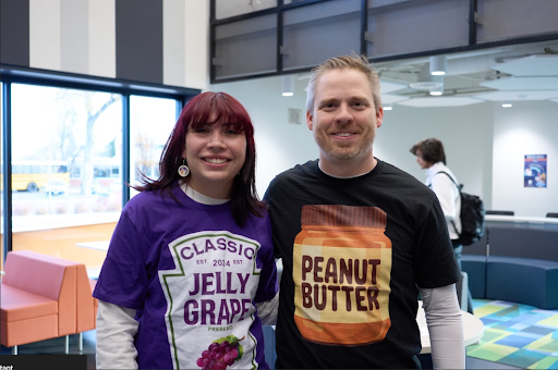 A young woman wearing a "Jelly Grape" shirt and a man wearing a "Peanut Butter" shirt smiling together in a brightly lit, modern common area.