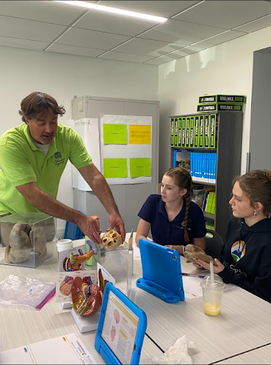 A male instructor in a green shirt demonstrating an anatomical model to two students sitting at a table with other models and tablets.