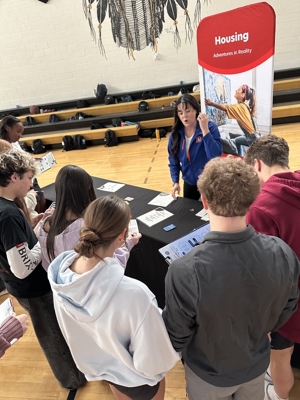 A group of students gathered around a table with a "Housing Adventures in Reality" banner in a gym.