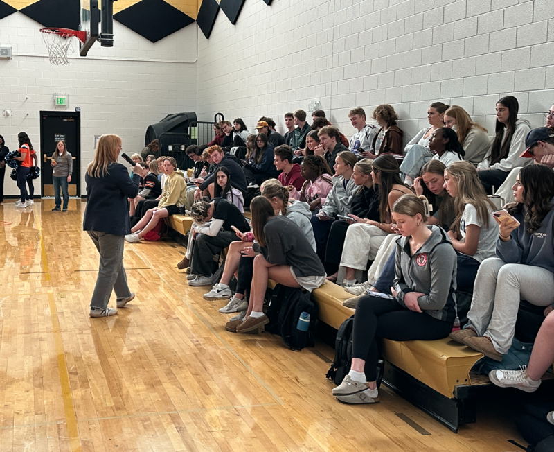 Students seated on bleachers in a gym, listening to a presentation by an adult woman walking in front of them.
