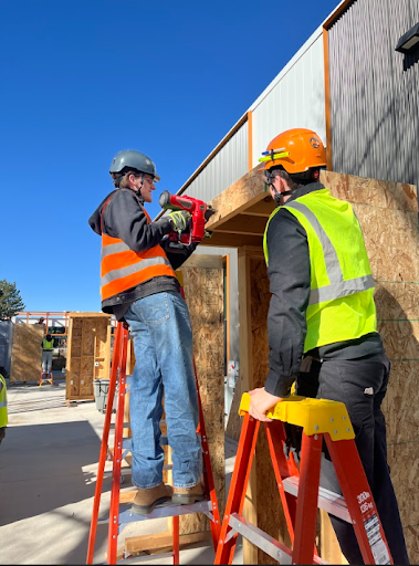Two construction workers on ladders framing a small structure, one using a pneumatic nail gun while the other spots.