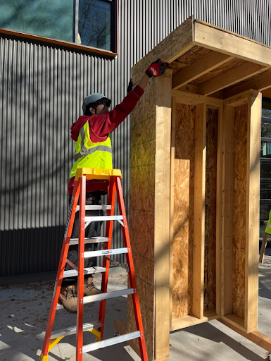 Construction worker on an A-frame ladder installing OSB sheathing on a wood-framed structure outside a modern building.