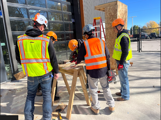 A group of five construction workers wearing safety vests and hard hats cutting wood on saw horses outdoors.