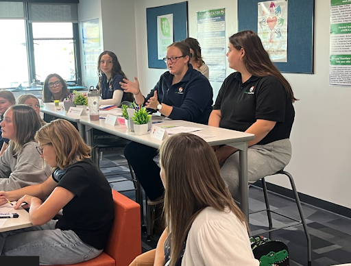 A panel of two female educators speak to a group of seated students during a classroom session.