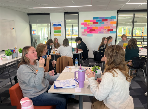 Students and adult educators collaborate and chat in small groups around tables in a brightly lit modern classroom.