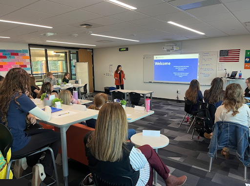An educator presents a "Welcome" slide on a screen to a room full of students seated at modern tables and chairs.