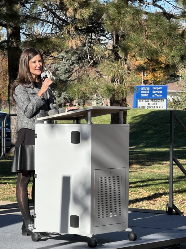 A woman speaks at a white podium outdoors on a sunny day. A blue sign is visible behind her.