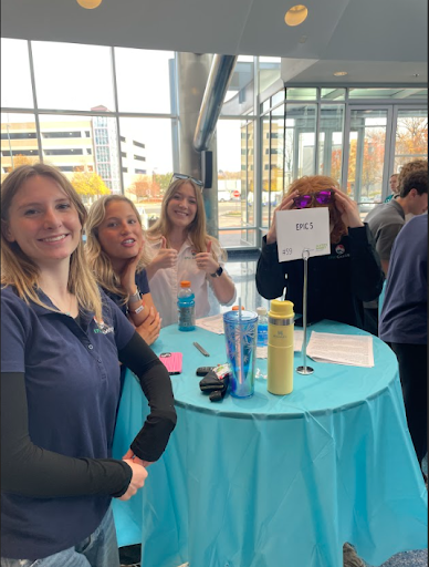 Four young women smiling and giving thumbs up while standing around a blue-covered table labeled "EPIC 5" at an indoor event.