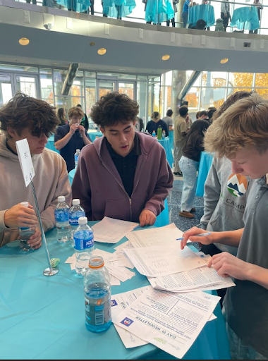 Three young men, possibly high school students, are reviewing paperwork and documents spread out on a blue-covered table at an event.