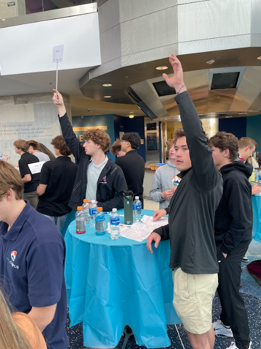 Two young men raising their hands in a crowded atrium, one holding up a small white sign, possibly participating in an auction or Q&A.