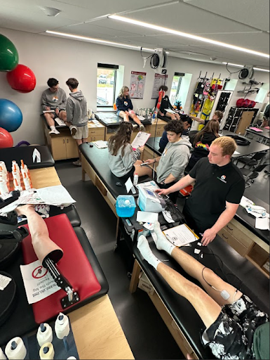 A dynamic scene in a sports medicine lab where students are practicing with equipment like stimulators and prosthetic limbs on examination tables.