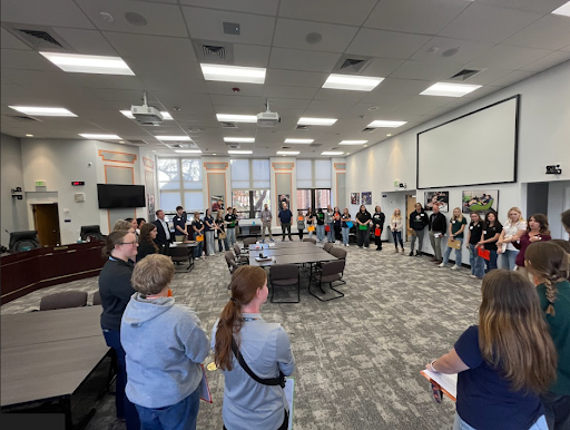 A large group of people, including young women and adults, standing in a large conference room forming a circle around a central table.