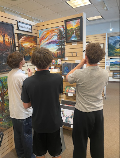 Three young men standing inside an art gallery, looking closely at various paintings on display.
