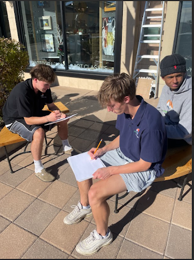 Three young men, two writing on clipboards, sitting outside on a bench in front of a storefront on a sunny day.