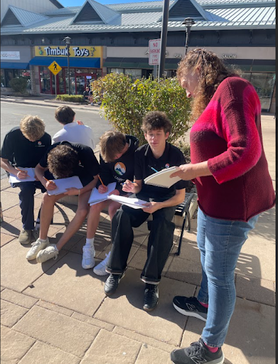A group of male students sitting outside on a bench, writing, while a female teacher stands next to them giving instructions.