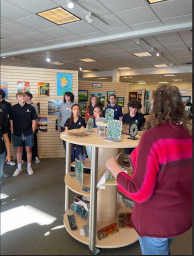 A group of students in a retail space, likely an art gallery, standing around a display as a woman addresses them.