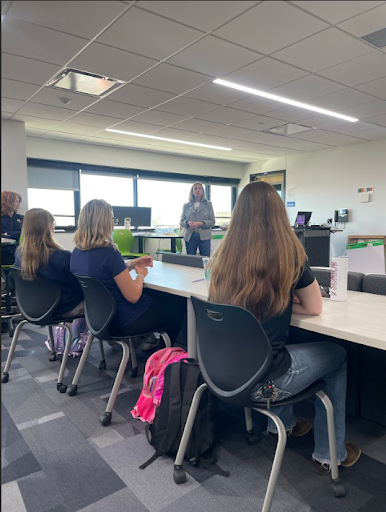 A classroom setting where a woman is standing and giving a presentation to three students seated facing away from the camera.