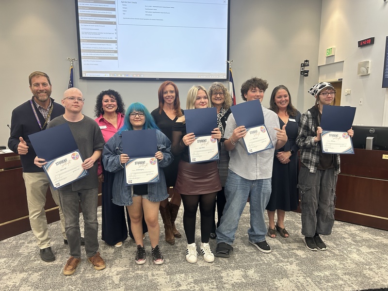 Students and adults pose holding "Student Spotlight" certificates at a school board meeting.