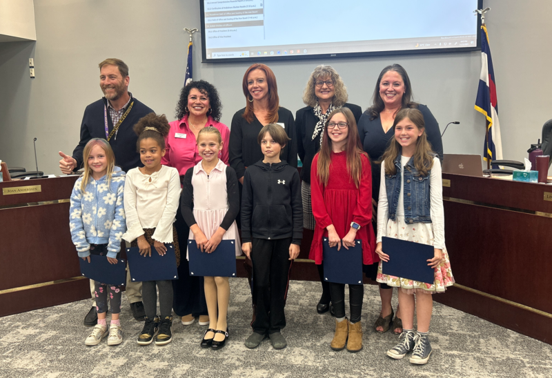 Elementary students and adults smile while holding certificates after being recognized at a board meeting.