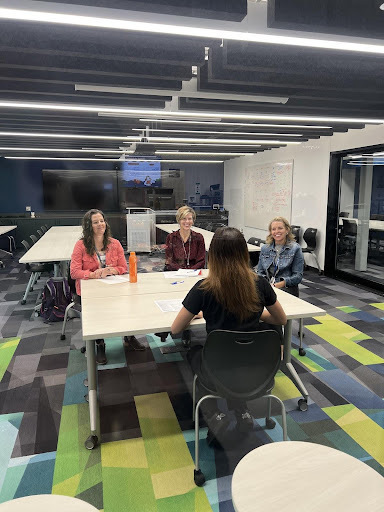A person with long hair sits at a table facing three smiling women across from them. They are in a modern meeting room with acoustic panels on the ceiling and a colorful patterned carpet.
