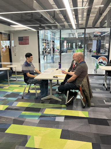 A young man in a collared shirt sits at a table, smiling and talking to two older men. They appear to be in an interview or meeting in a bright, modern office space with a colorful carpet.