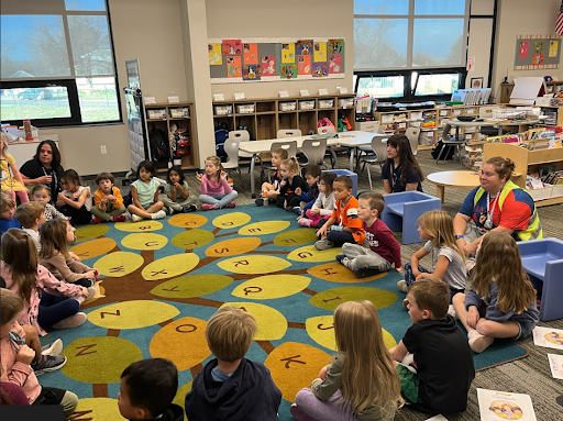 A large group of young children and two adults sit in a circle on a colorful classroom rug decorated with a tree and letters, participating in a story or group time activity.