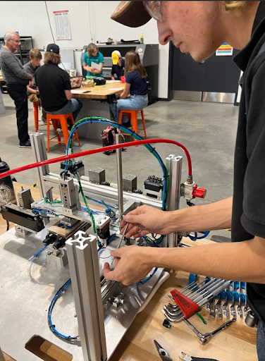 A young man works on a small machine, using a tool to adjust a component. Tools are laid out next to him, and a group of people is visible in the background.