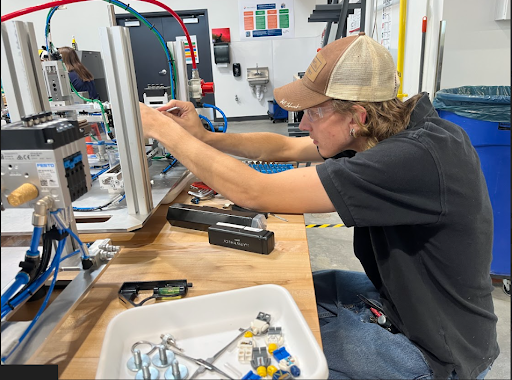 A young man in a cap and safety glasses leans over a workbench, focusing on adjusting a component on a piece of machinery. A container of parts is in the foreground.