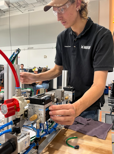 A young man wearing safety glasses and a black shirt adjusts a pneumatic control panel with blue and red tubing in a modern training workshop.