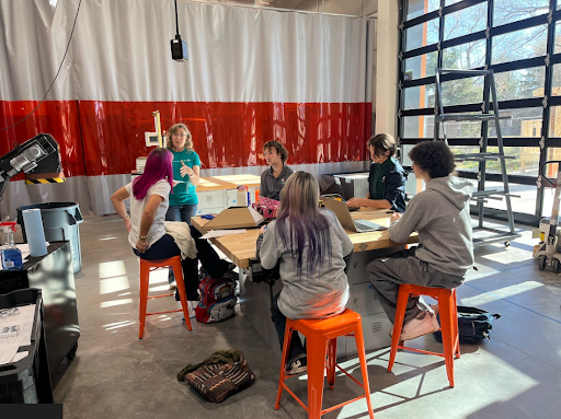 Five students and one instructor gather around a workbench in a bright workshop space, engaged in a discussion. A large glass garage door is visible on the right.