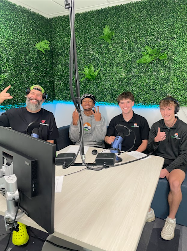 Three young men and one older man with a beard are sitting at a table with microphones in a podcast studio, smiling and making hand gestures.